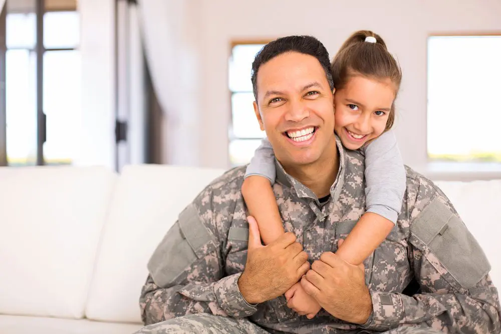 A young military father smiles at the camera with his daughter, who has her arms around his shoulders