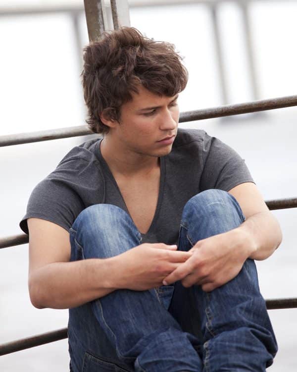 Teenage boy sitting with his arms around his knees, frowning and appearing to look toward the ground