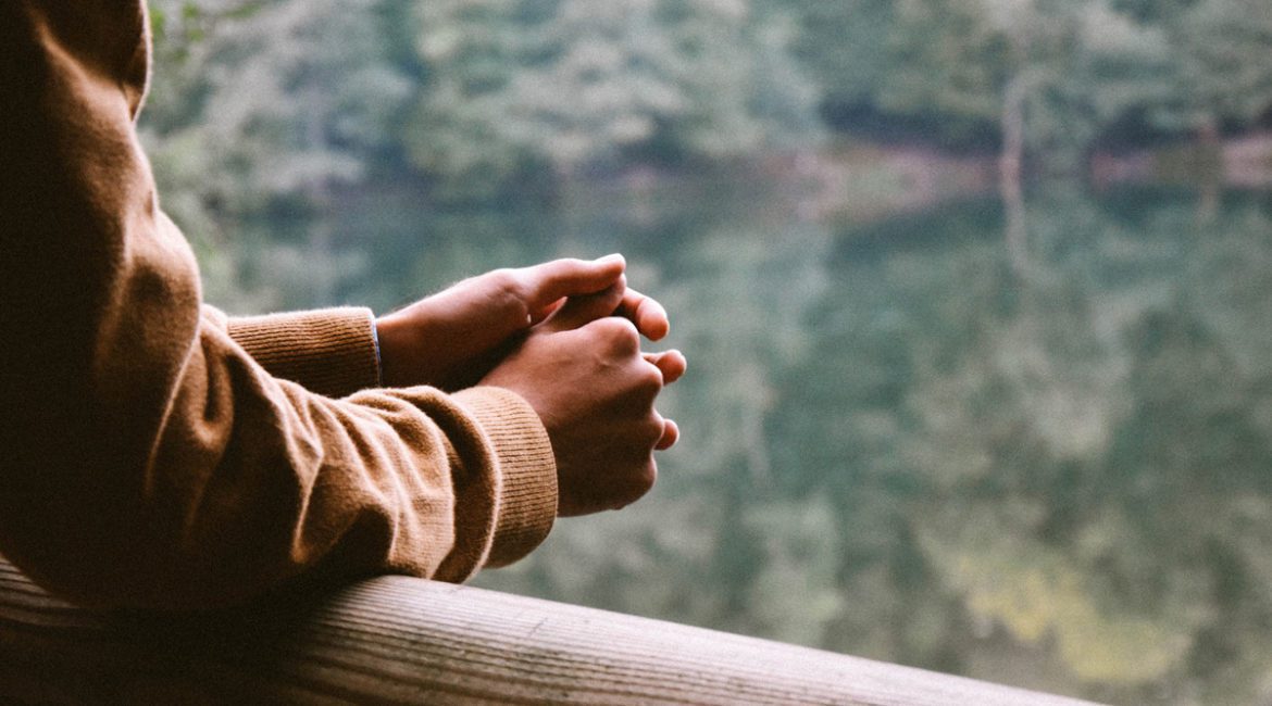 A closeup of a person's hands nervously clasped together as they look out over a river in a wooded area