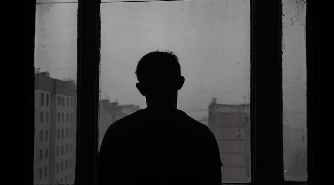 A black and white photo of a man's silhouette as he looks out a window at an urban landscape.