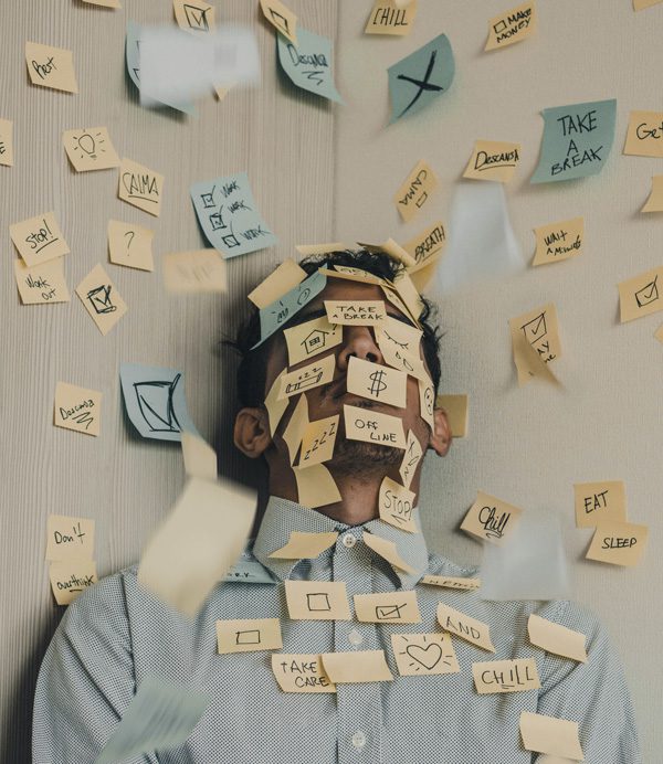 A stressed man with a variety of sticky notes on his face, body, and all over the wall around him