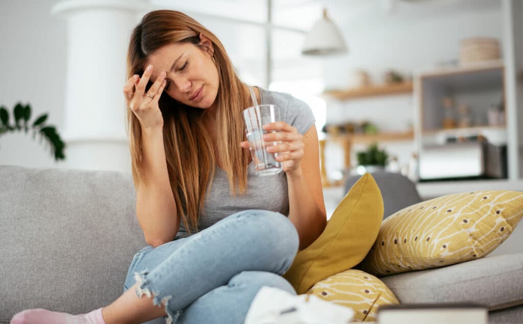 A sick woman holding her head and glass of water while sitting on a couch